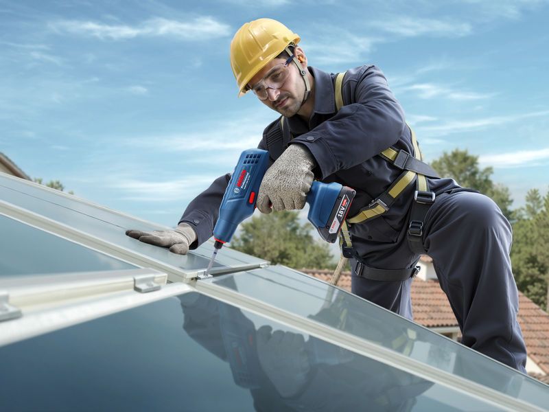 Một người mặc đồ bảo hộ đang siết chặt bu lông trên tấm pin mặt trời trên mái nhà bằng dụng cụ điện.