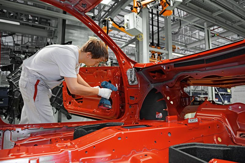 Một người mặc đồ bảo hộ đang sử dụng máy khoan không dây để lắp ráp cửa ô tô trong nhà máy.