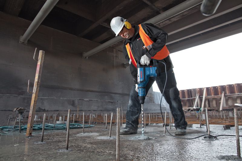 Một người mặc đồ bảo hộ đang khoan bê tông bằng búa quay tại một công trường xây dựng.