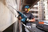 Một người mặc đồ bảo hộ đang khoan vào bức tường bê tông tại một công trường xây dựng.