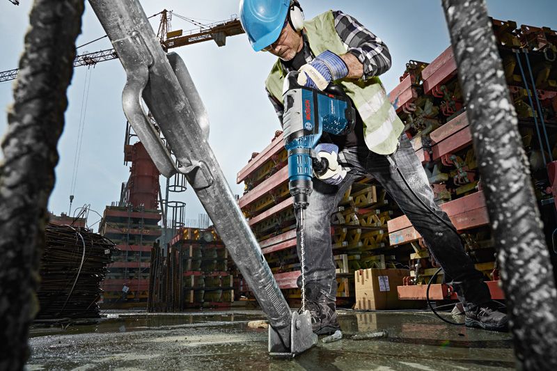 GBH 8-45 D Một công nhân mặc đồ bảo hộ đang sử dụng máy khoan búa để khoan vào nền bê tông tại một công trường xây dựng.