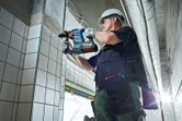 Một người mặc đồ bảo hộ đang khoan vào tường lát gạch bằng máy khoan không dây.