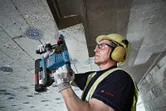 Một người mặc đồ bảo hộ đang khoan trần bê tông bằng búa quay không dây.