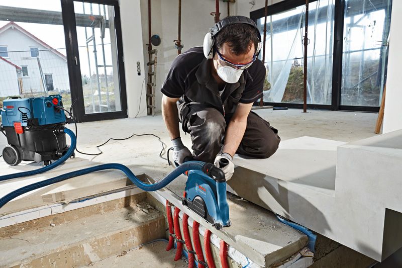 Một người đàn ông mặc đồ bảo hộ đang dùng máy đục tường để cắt bê tông gần các đường ống lộ thiên.