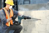 Một người mặc đồ bảo hộ đang sử dụng búa quay để khoan vào tường bê tông.