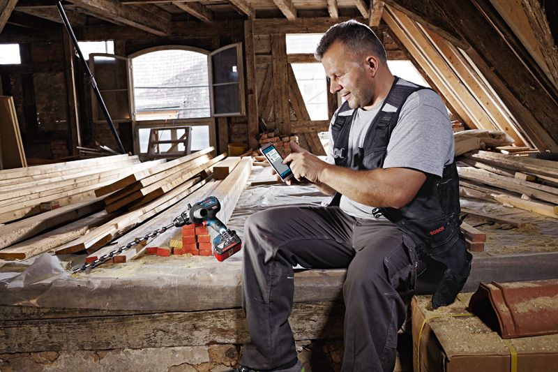 Một người đeo thiết bị an toàn đang sử dụng điện thoại thông minh bên cạnh máy khoan không dây trong xưởng mộc.