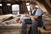 Một người đeo thiết bị an toàn đang sử dụng điện thoại thông minh bên cạnh máy khoan không dây trong xưởng mộc.