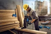 Một người mặc đồ bảo hộ đang khoan vào thanh dầm gỗ tại một công trường xây dựng.