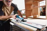 Một người mặc đồ bảo hộ đang sử dụng máy cưa tròn không dây để cắt gỗ trong xưởng.