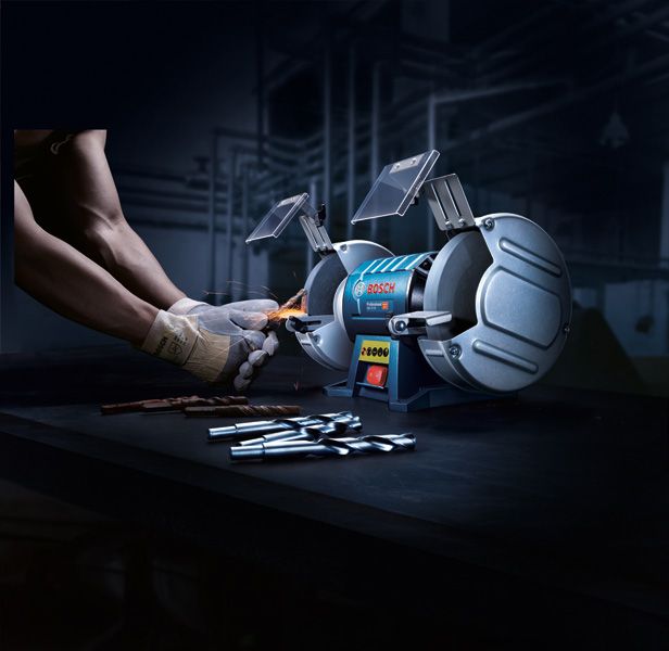 Một người đeo thiết bị an toàn đang mài mũi khoan bằng máy mài bàn có hai bánh xe.