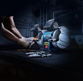 Một người mặc đồ bảo hộ đang mài mũi khoan bằng máy mài bàn có hai bánh xe.
