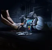 Một người đeo thiết bị an toàn đang mài mũi khoan bằng máy mài bàn có hai bánh xe.