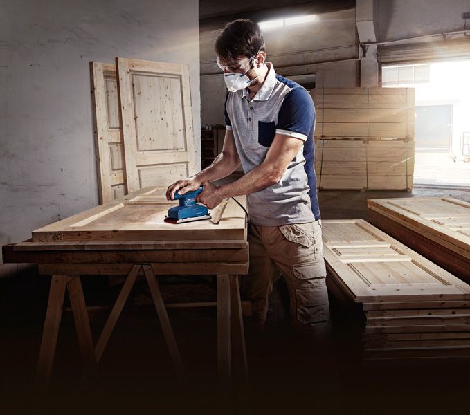 Một người mặc đồ bảo hộ đang chà nhám cửa gỗ bằng máy chà nhám quỹ đạo trong xưởng.