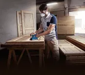 Một người mặc đồ bảo hộ đang chà nhám cửa gỗ bằng máy chà nhám quỹ đạo trong xưởng.