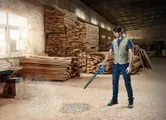 Một người đàn ông mặc đồ bảo hộ đang dùng máy thổi để thổi sạch mùn cưa trong xưởng mộc.