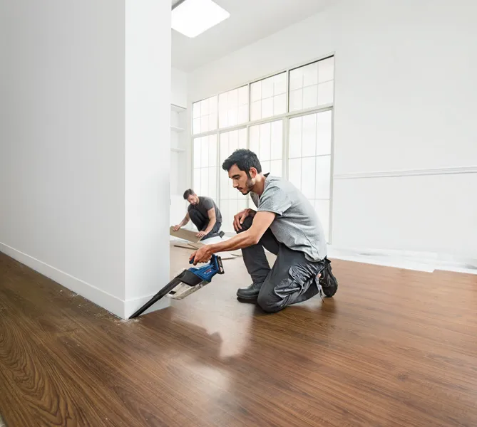 GAS 18V-1 Một người đang cắt sàn gần tường bằng máy cưa không dây trong khi một người khác đang lắp tấm sàn.