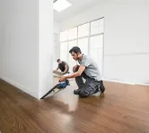 Một người đang cắt sàn gần tường bằng máy cưa không dây trong khi một người khác đang lắp tấm sàn.