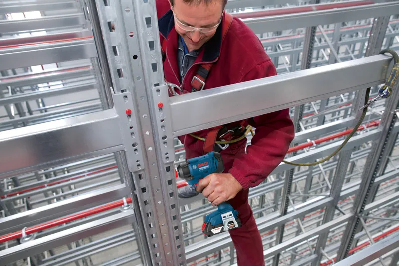 Một người mặc đồ bảo hộ đang sử dụng máy khoan điện trên thanh dầm kim loại trong một nhà kho.