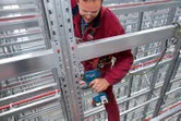 Một người mặc đồ bảo hộ đang sử dụng máy khoan điện trên thanh dầm kim loại trong một nhà kho.