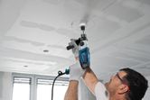 Một người mặc đồ bảo hộ đang khoan một lỗ tròn trên trần nhà bằng một dụng cụ điện.