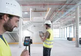 Hai người mặc thiết bị an toàn sử dụng máy đo laser và máy tính bảng bên trong một công trường xây dựng lớn.