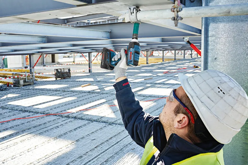 Một người mặc thiết bị an toàn sử dụng cờ lê tác động không dây trên một thanh kim loại.