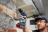 Một người mặc đồ bảo hộ đang khoan vào tường gạch bằng máy khoan điện không dây.