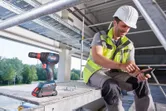 Một người mặc đồ bảo hộ đang sử dụng điện thoại thông minh bên cạnh máy khoan không dây tại một công trường xây dựng.
