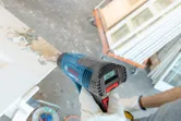 Một người mặc đồ bảo hộ đang sử dụng súng nhiệt để cạo lớp sơn trên cửa.
