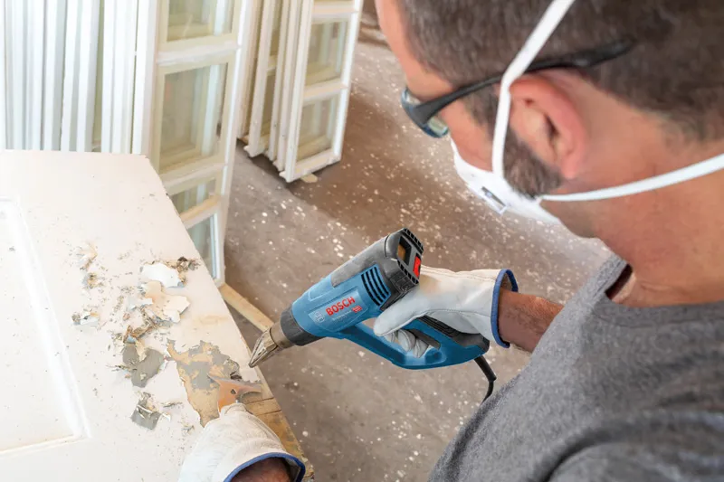 Người mặc thiết bị an toàn sử dụng súng nhiệt để cạo lớp sơn trên cửa.