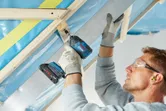 Một người mặc đồ bảo hộ đang dùng máy khoan điện để cố định một thanh kim loại vào khung gỗ.