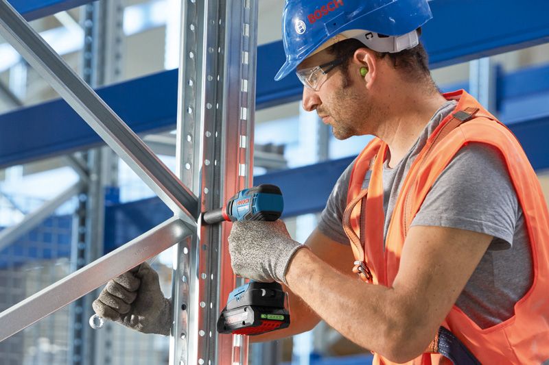 Một người mặc đồ bảo hộ sẽ cố định khung kim loại bằng máy khoan điện không dây.