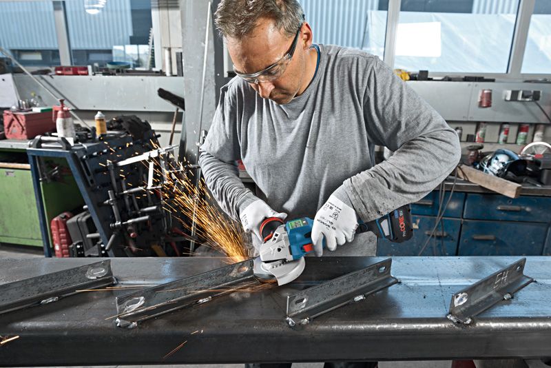 Một người mặc đồ bảo hộ đang sử dụng máy mài góc để cắt một thanh kim loại.