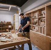 Một người mặc đồ bảo hộ đang sử dụng máy khoan không dây trên một chiếc hộp gỗ trong xưởng.