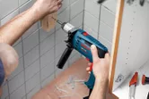 Một người đang khoan vít vào khối gỗ gắn trên bức tường lát gạch.