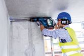Một người mặc đồ bảo hộ đang khoan vào trần bê tông bằng búa quay.