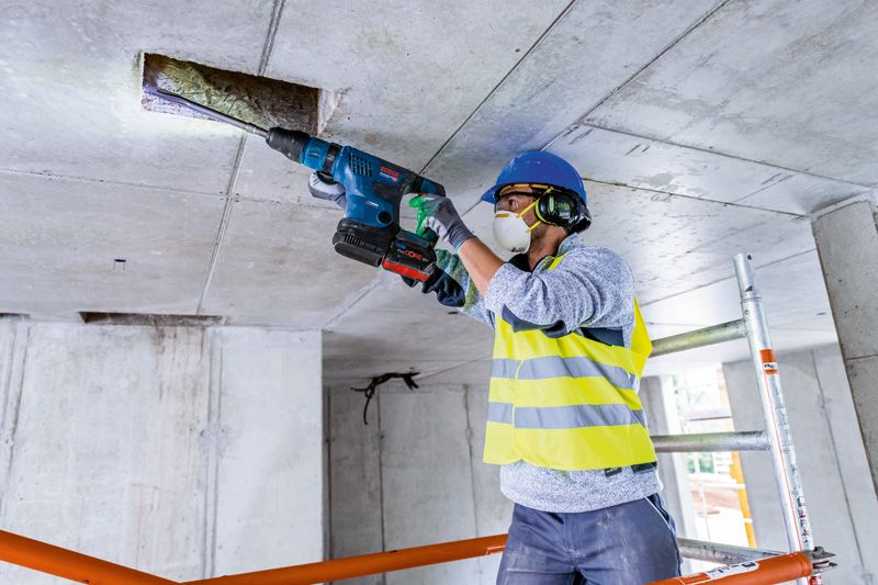 Một người mặc đồ bảo hộ đang khoan trần bê tông bằng búa quay không dây.