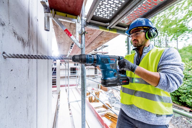 Một người đàn ông mặc đồ bảo hộ đang dùng máy khoan búa xoay không dây để khoan vào bê tông.