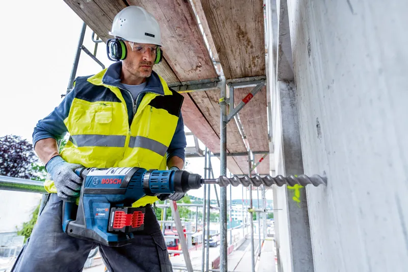 Một người đàn ông mặc đồ bảo hộ đang dùng máy khoan búa xoay không dây để khoan vào bê tông.