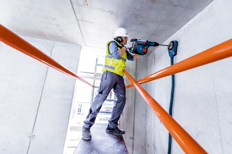 Một người mặc đồ bảo hộ đang khoan vào trần bê tông bằng búa quay.