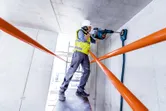 Một người mặc đồ bảo hộ đang khoan vào trần bê tông bằng búa quay.