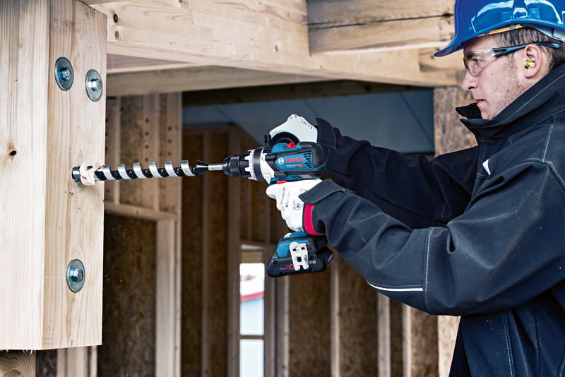 Một người mặc đồ bảo hộ đang khoan vào thanh dầm gỗ tại một công trường xây dựng.