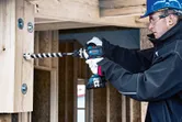 Một người mặc đồ bảo hộ đang khoan vào thanh dầm gỗ tại một công trường xây dựng.