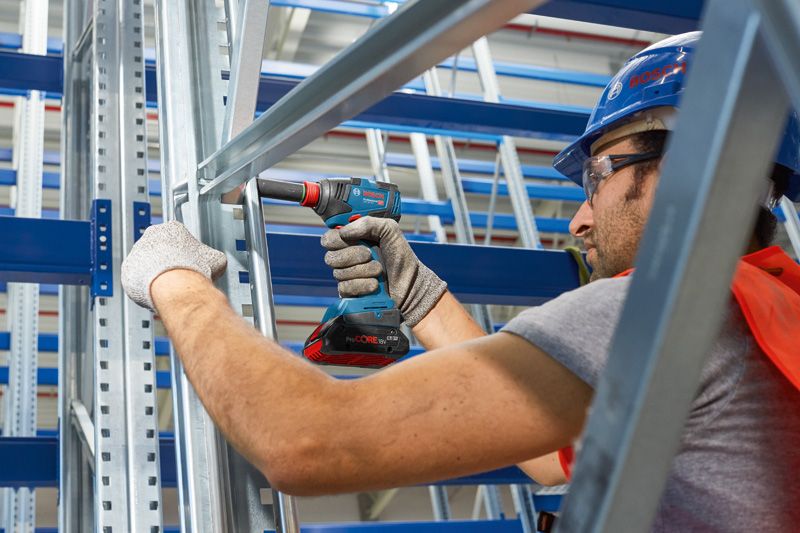 Một người mặc đồ bảo hộ đang sử dụng máy khoan không dây trên khung kim loại.