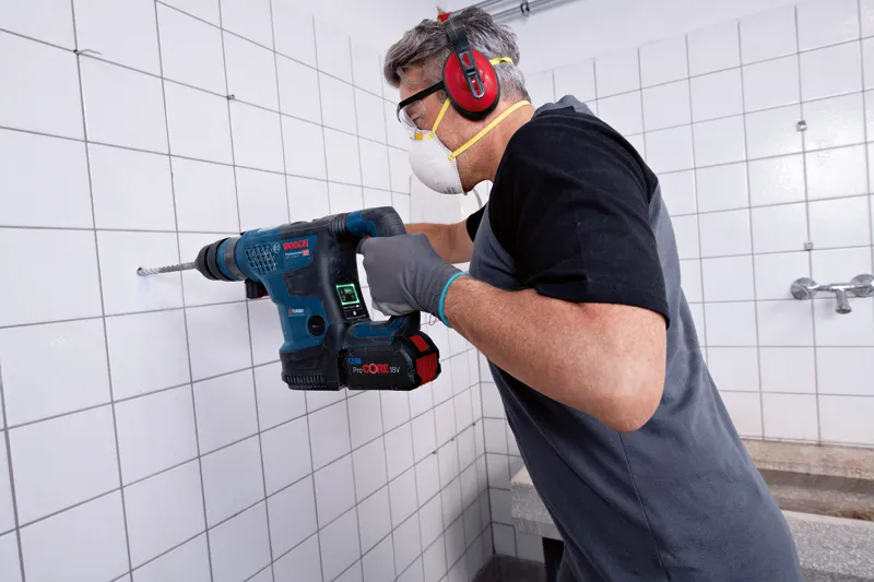 GBH 18V-34 CF Một người mặc đồ bảo hộ đang khoan vào bức tường lát gạch bằng máy khoan không dây.
