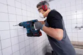 Một người mặc đồ bảo hộ đang khoan vào bức tường lát gạch bằng máy khoan không dây.