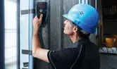 Một người mặc thiết bị an toàn đang quét bức tường bê tông bằng một công cụ dò tìm.
