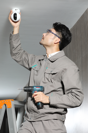 Một người đàn ông mặc đồ bảo hộ đang lắp đặt camera an ninh bằng máy khoan.