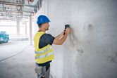 Một người mặc đồ bảo hộ đang quét bức tường bê tông bằng thiết bị dò tìm.