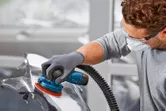 Một người mặc đồ bảo hộ đang chà nhám một bộ phận của ô tô bằng máy chà nhám quỹ đạo ngẫu nhiên không dây.